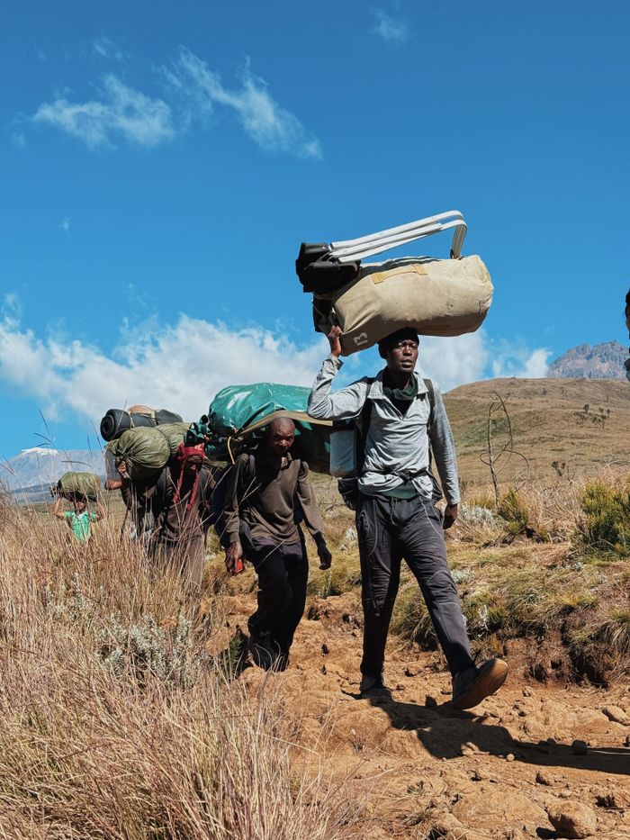 Group of African porters carrying gear on a mountainous trail under a sunny blue sky.
